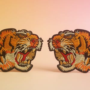 Puede incluir: Dos parches bordados con cabezas de tigre. Los parches presentan hilos de color naranja, negro y crema. Los tigres tienen la boca abierta con el interior rojo, mostrando los dientes. Los parches están sobre un fondo degradado de melocotón y rosa.