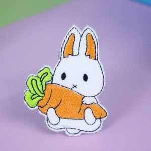 May include: White embroidered bunny holding a large orange carrot with green leafy tops.