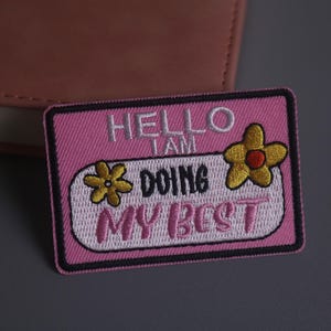 以下が含まれることがあります： 「HELLO I AM DOING MY BEST」の文字が白とピンクの糸で刺繍されたピンク色のワッペン。赤い中心を持つ2つの黄色の花がワッペンに刺繍されています。ワッペンは黒い縁取りがあります。