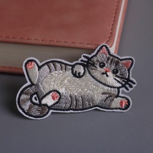 May include: Embroidered patch of a gray and white cat lying on its back. The cat has pink accents on its paws and ears, with black whiskers and a black outline. The patch is on a gray surface.
