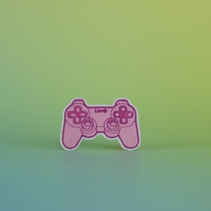 May include: Pink embroidered video game controller patch with white stitching. The controller has a pink background and white buttons. The text "Love" is embroidered in white on the controller.