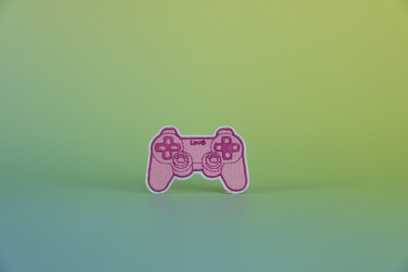 May include: Pink embroidered video game controller patch with white stitching. The controller has a pink background and white buttons. The text "Love" is embroidered in white on the controller.