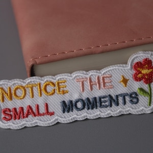 Puede incluir: Parche blanco bordado con el texto "NOTICE THE SMALL MOMENTS" en letras de colores. Una flor roja con centro amarillo está bordada en el lado derecho del parche.
