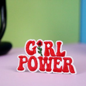 Pode incluir: Etiqueta bordada vermelha com as palavras "GIRL POWER" e uma pequena flor vermelha.
