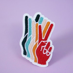 May include: Embroidered patch featuring a hand making the peace sign. The patch has a white border and a layered design with colors including red, orange, yellow, tan, teal, and black. The background is a solid lavender color.