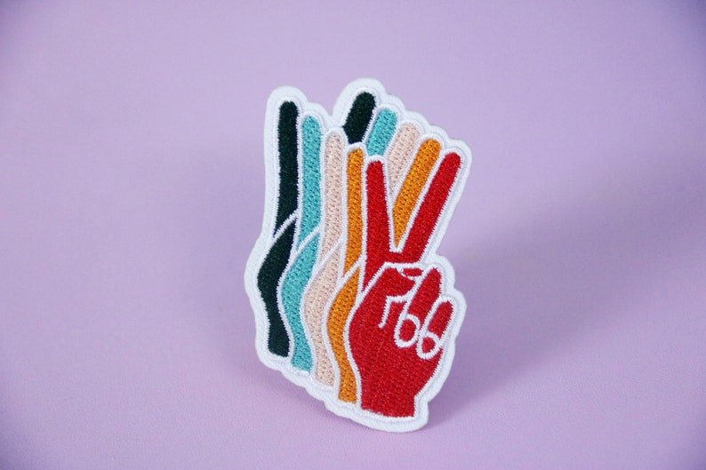 Pu&ograve; includere: Toppa ricamata raffigurante una mano che fa il segno della pace. La toppa ha un bordo bianco e un design a strati con colori tra cui rosso, arancione, giallo, beige, turchese e nero. Lo sfondo &egrave; di colore lavanda.
