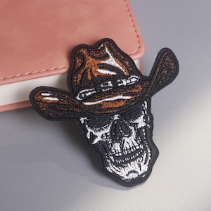 Può includere: Toppa ricamata raffigurante un teschio con un cappello da cowboy. Il cappello è marrone e bianco, mentre il teschio è nero e bianco. La toppa è destinata a vestiti o accessori.