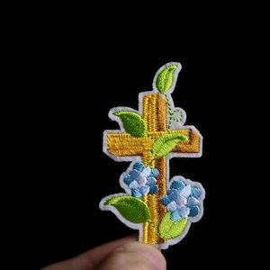 以下が含まれることがあります： 緑の葉と青い花で飾られた金の十字架が特徴の刺繍パッチ。 パッチは白い縁取りがあり、黒い背景に配置されています。 衣類やアクセサリーに最適です。