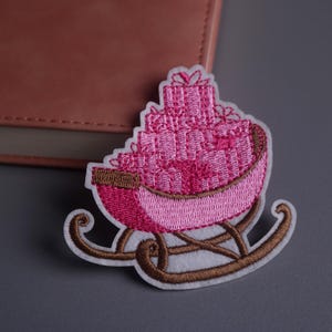 Könnte beinhalten: Gestickter Aufnäher mit einem rosa Schlitten voller Geschenke. Der Schlitten hat braune Kufen und einen weißen Rand. Die Geschenke sind hoch gestapelt und mit rosa Nähten versehen. Ein charmantes, festliches Design.