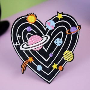Könnte beinhalten: Ein schwarzer herzförmiger Patch mit einem gestickten Design einer Galaxie mit Planeten, Sternen und einem Kometen. Die Planeten sind rosa, gelb, blau und lila.