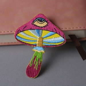 Op de afbeelding: Een geborduurde paddenstoel patch in roze en geel met een blauw oog bovenop. De paddenstoel heeft een groene steel en witte strepen op de dop.