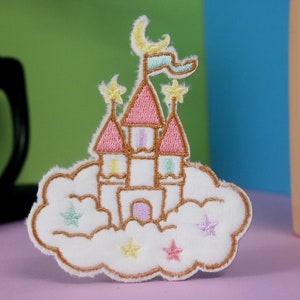 Könnte beinhalten: Ein weißer gestickter Aufnäher mit einem rosa Schloss auf einer Wolke mit Sternen. Das Schloss hat einen Halbmond und eine Flagge oben drauf.