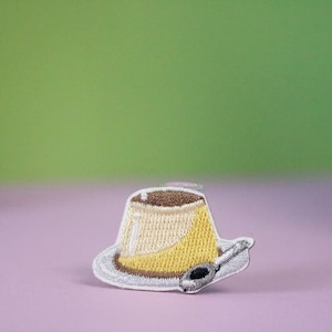 May include: A yellow and brown embroidered patch of a pudding dessert with a spoon on a white plate.
