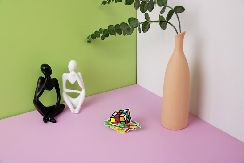 May include: Two abstract sculptures of people sitting on a pink surface, a vase with a leafy green plant, and a colorful sticker of a melting Rubik's cube.