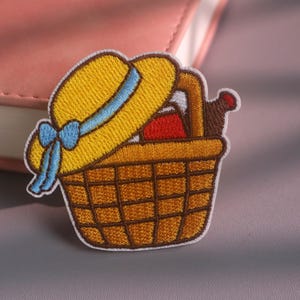 Puede incluir: Un parche bordado que muestra una cesta de picnic tejida de color marrón llena con un objeto rojo y una botella marrón oscuro, rematada con un sombrero de paja amarillo con una cinta azul claro y un lazo, todo bordeado con hilo blanco.