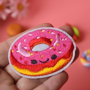 Puede incluir: Parche de donut bordado de color rosa con glaseado blanco y chispas. El donut tiene una cara feliz y está rodeado de un borde blanco.