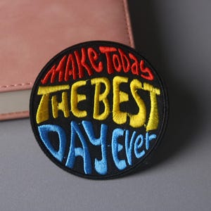 May include: A round, embroidered patch with the phrase "Make Today The Best Day Ever" in colorful, stylized lettering. The text is in red, yellow, and blue against a black background. The patch is on a gray surface.