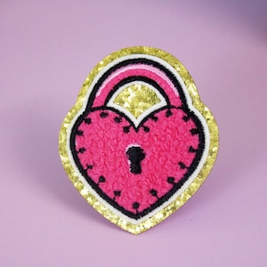 Puede incluir: Un parche en forma de corazón con un diseño de cerradura. El parche presenta un centro rosa texturizado, detalles en blanco y negro y un borde dorado con purpurina. La cerradura tiene un ojo de cerradura y un contorno negro.