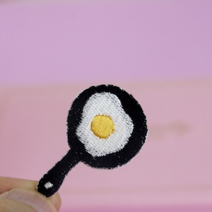 May include: A black embroidered iron-on patch of a frying pan with a white fried egg inside. The yolk of the egg is yellow.