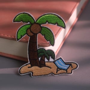 Puede incluir: Un parche bordado que muestra una escena de isla tropical con dos palmeras marrones de hojas verdes, un área de arena color canela y una pequeña tumbona a rayas azules y blancas. El parche tiene un borde blanco y costuras detalladas.