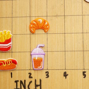 May include: A set of five iron-on patches featuring various food items, including a croissant, french fries, a hot dog, and a glass of orange juice. The patches are arranged on a gridded surface with inch markings.