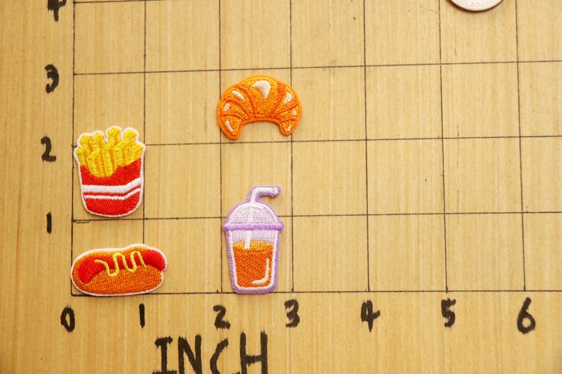 May include: A set of five iron-on patches featuring various food items, including a croissant, french fries, a hot dog, and a glass of orange juice. The patches are arranged on a gridded surface with inch markings.
