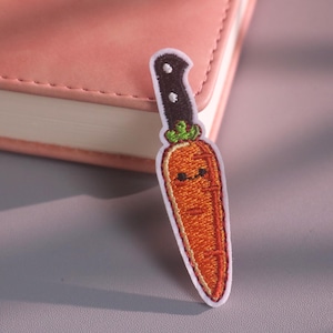 Op de afbeelding: Een geborduurde patch met een feloranje wortel met een lachend gezicht en een groen blad, bevestigd aan een zwart mesheft. Het ontwerp is omlijnd met witte draad, wat een duidelijke vorm creëert die een groente en een mes combineert.