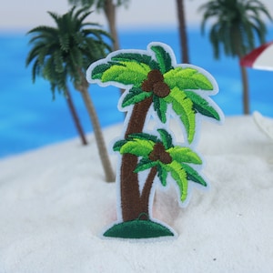 Puede incluir: Parche bordado de una palmera verde con tronco marrón y cocos. El parche está sobre una superficie de arena blanca con una escena de playa en miniatura.