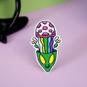 May include: Embroidered iron-on patch featuring a green alien with a pink and white mushroom growing out of its head. The mushroom has a rainbow-colored stem.