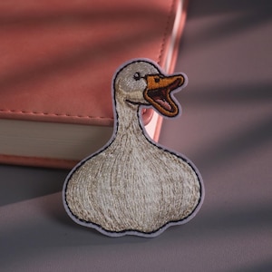 May include: An embroidered patch featuring a white goose with a long neck and an open orange beak. The goose has detailed stitching in shades of white, gray, and brown, with a dark eye and a distinct outline, creating a textured appearance.