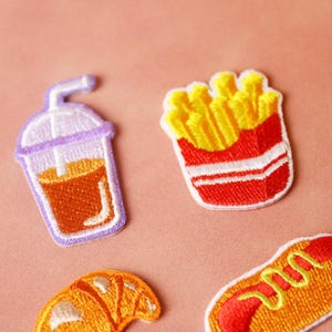 May include: Four embroidered iron-on patches featuring a glass of iced tea, a carton of french fries, a croissant, and a hot dog. The patches are on a pink background.