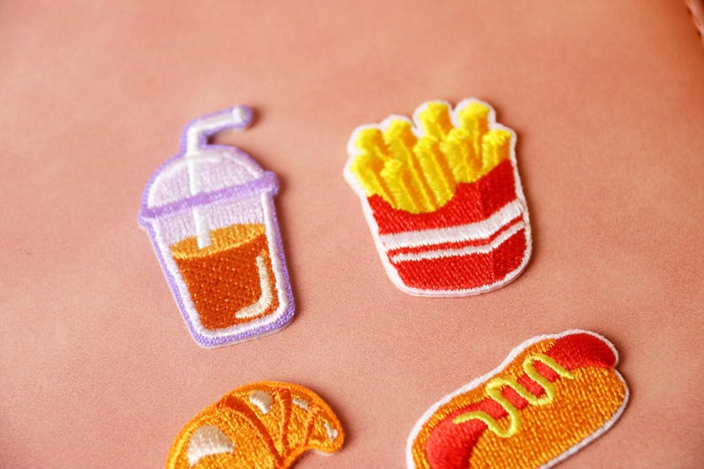 May include: Four embroidered iron-on patches featuring a glass of iced tea, a carton of french fries, a croissant, and a hot dog. The patches are on a pink background.