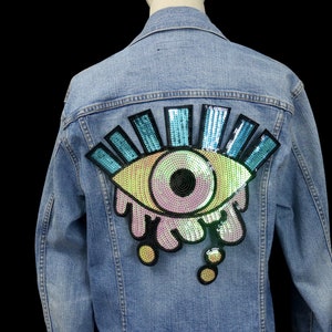 Può includere: Una giacca di jeans blu chiaro con un grande patch a forma di occhio in paillettes iridescenti sul retro. L'occhio è contornato di nero e ha un'iride rosa e una pupilla nera. L'occhio è circondato da paillettes blu e verdi.