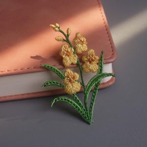 May include: Embroidered floral brooch featuring yellow flowers and green leaves. The brooch is placed on a pink leather-bound notebook, with a gray surface in the background. The brooch is a decorative accessory.