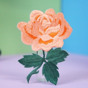 May include: Peach-colored embroidered rose with green leaves. The rose is a single bloom with a stem and leaves. The rose is a popular motif for embroidery and is often used in floral designs.