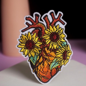 Puede incluir: Parche bordado con un diseño de corazón humano estilizado con girasoles. El corazón está representado en tonos naranja y rojo, con girasoles amarillos y hojas verdes. El parche tiene un borde blanco.