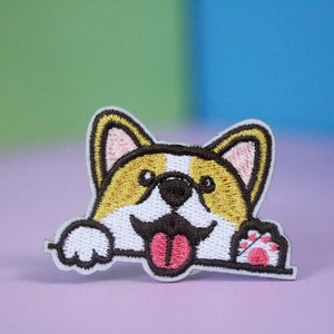 Könnte beinhalten: Gestickter Aufnäher eines Cartoon-Corgis mit braunen Ohren, einem weißen Gesicht und einer herausgestreckten rosa Zunge. Der Corgi schaut über eine weiße Oberfläche mit erhobenem Pfote.