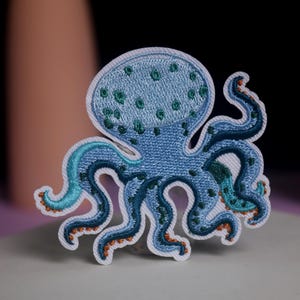 Könnte beinhalten: Gestickter Oktopus-Aufnäher in Blau-, Türkis- und Orangetönen. Der Oktopus hat einen hellblauen Kopf mit grünen Punkten und dunkelblauen Tentakeln. Der Aufnäher ist weiß umrandet.