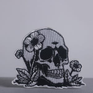 May include: Embroidered patch featuring a skull design with flowers. The skull is white with black details, including the eye sockets, nose, and teeth. Black flowers and leaves surround the skull, creating a contrasting design.