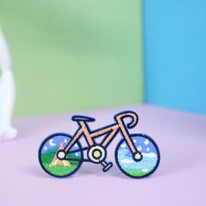 May include: An embroidered iron-on patch of a bicycle with a mountain scene in one wheel and a sky scene in the other wheel, in blue and brown.