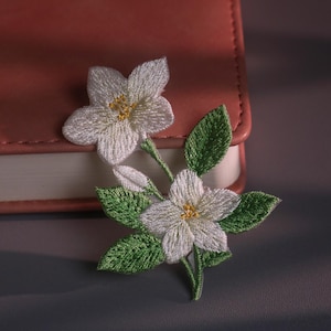 May include: Embroidered white flower with yellow centres and green leaves. The floral design is set against a pink leather-bound notebook. The delicate embroidery showcases the intricate details of the flower and leaves.