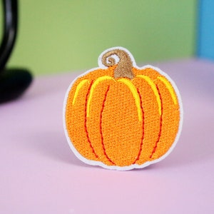 May include: Orange embroidered pumpkin patch iron-on applique. The pumpkin is outlined in white and has yellow stitching for the stem and lines.