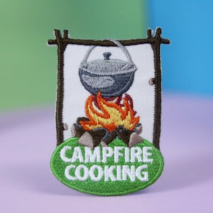 Könnte beinhalten: Ein gestickter Aufnäher mit einem grünen ovalen Hintergrund und dem Text "Campfire Cooking". Der Aufnäher zeigt ein Lagerfeuer mit einem Topf, der darüber hängt.