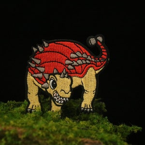 May include: Embroidered patch of a cartoon Ankylosaurus dinosaur. The patch features a red and yellow colour scheme with grey spikes along its back. The dinosaur has a friendly expression with a wide grin. The patch is on a green, mossy surface.