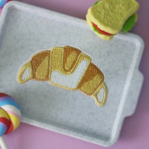 May include: A white ceramic tray with a brown and yellow embroidered croissant patch. The tray is surrounded by colourful lollipops, a sandwich, and a hot dog.
