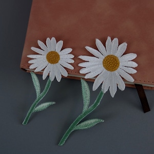 Puede incluir: Dos parches de hierro de flores de margarita blancas con tallos y hojas verdes sobre un diario de cuero rosa.