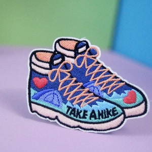 以下が含まれることがあります： 青いハイキングブーツのアイロン接着パッチ。オレンジ色の靴紐とブーツの底に「TAKE A HIKE」の文字が刺繍されています。