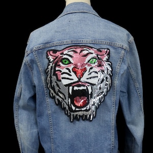 May include: Light blue denim jacket featuring a large, sequined tiger face on the back. The tiger has pink, silver, and red accents with green eyes. The jacket is a classic style with a collar and button closures.