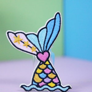 May include: A colorful embroidered iron-on patch featuring a mermaid tail with a pink, blue, and yellow scale pattern, a pink heart, and gold stars. The tail is surrounded by a blue wave.