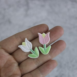 Pode incluir: Dois patches bordados para passar a ferro com forma de tulipas. Um é branco com um centro amarelo e o outro é rosa com um centro amarelo.